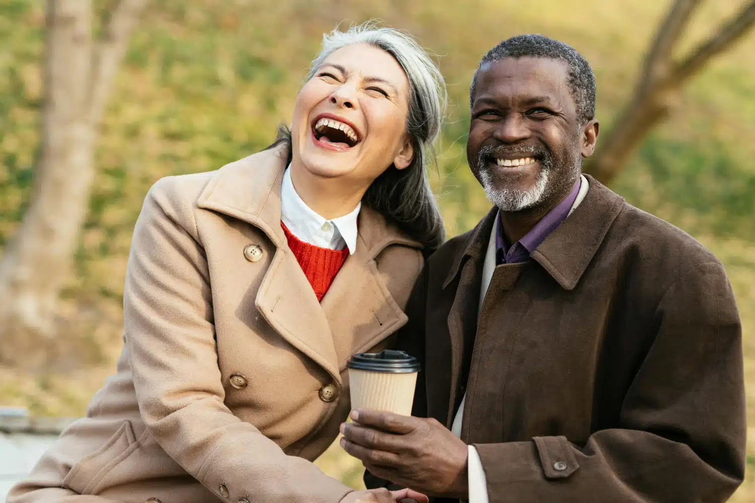 Older asian woman wearing a tan coat and laughing with an african- american man in a brown coat holding coffee in a park