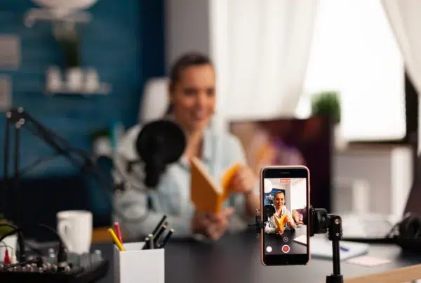 Smartphone on a tripod recording a woman filming a talking-head video at her desk with a microphone and softbox lighting.