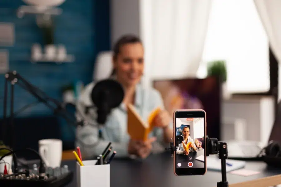Smartphone on a tripod recording a woman filming a talking-head video at her desk with a microphone and softbox lighting.
