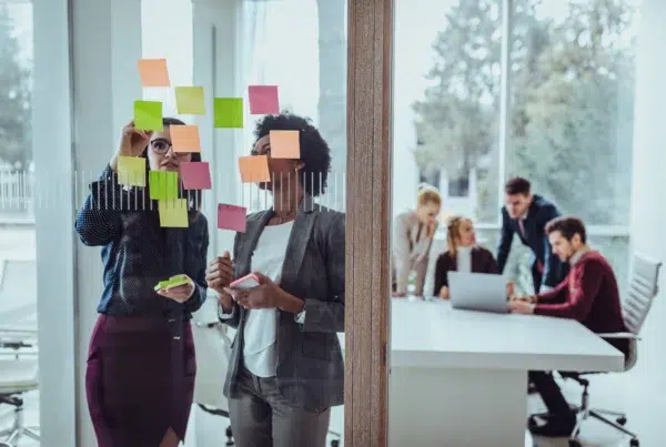 Two businesswomen collaborate on a glass wall covered in colorful sticky notes, brainstorming ideas while holding additional notes, with a group of coworkers working at a laptop in a meeting room in the background.