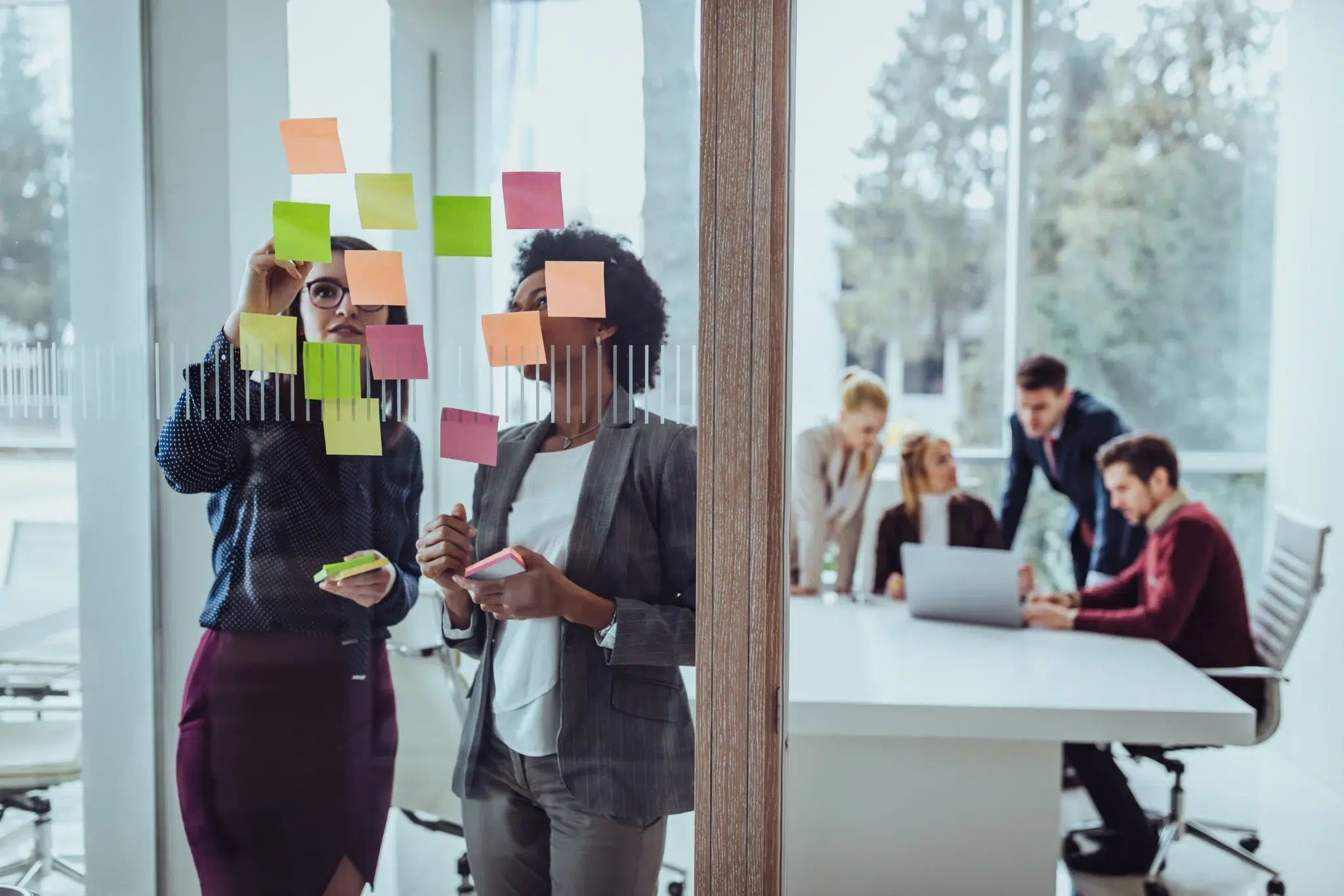 Two businesswomen collaborate on a glass wall covered in colorful sticky notes, brainstorming ideas while holding additional notes, with a group of coworkers working at a laptop in a meeting room in the background.